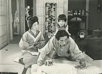 Une femme à genou (Kinuyo Tanaka) tient dans ses mains un porte-monnaie ouvert et regarde sa fille (Mitsuko Ichimura) assise à califourchon sur le dos de son père (Atsushi Watanabe) et qui s’agrippe à ses cheveux (image en noir et blanc).