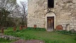 Photographie couleur de la façade d'un bâtiment en pierre de taille, dont l'entrée est fermée par une porte en bois. Au premier plan, des khatchkars sont disposés sur la pelouse.