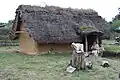 Photographie montrant un petit bâtiment couvert de chaume, avec une femme costumée à son entrée.