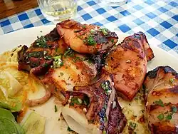 La photographie couleur présente des calmars farcis grillés et saupoudrés d'herbes. Ils sont posés sur un lit de salade, dans une assiette blanche sur une nappe à carreaux bleus.