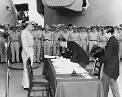 Photo d'un homme en train de signer un document sur une table. Il est entouré de soldats américains en train d'assister à la scène.