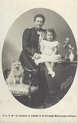 la comtesse de Flandre, portant une robe sobre et foncée, ainsi qu'un long sautoir sourit en tenant sa petite-fille en tenue claire, tandis qu'à droite est assis un loulou de Poméranie