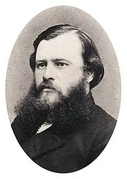 Portrait photographique en noir et blanc d'un trentenaire en costume, aux cheveux bruns mi-courts et à la barbe touffue.