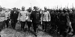 Photographie noir et blanc de trois hommes en uniforme paradant devant des troupes au garde à vous.