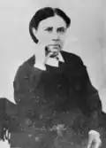 Dame assise, robe noire serrée par une ceinture, petit col blanc, poing sous le menton