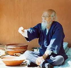 C'est une photographie en couleur de Fukuoka assis devant des récipients en céramique, il a un âge avancé.