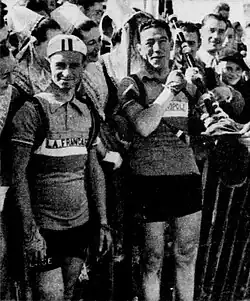 Photo en noir et blanc montrant deux hommes en tenue de cycliste se tenant devant des spectatrices portant des coiffes