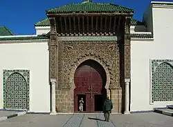Photographie d'une tour brune sombre ouverte d'une porte avec un arc outrepassé, richement décoré et couvert d'un toit vert (oxyde de cuivre ou céramique). L'ensemble est flanqué de deux murs blancs percés d'ouvertures plus simples.