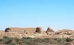 Des murs en ruine s'élèvent parmi de petits buissons épineux sous un ciel sans nuages ; dans une ouverture, on aperçoit une structure semblable à une mosquée, avec un chameau servant d'échelle de référence.