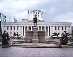Statue noire devant un bâtiment à colonnades dont le dôme est tronqué.