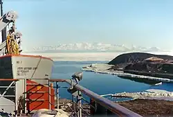 Proue d'un bateau vue depuis une plate-forme, avec une péninsule occupée par une cabane et, au-delà d'une mer couverte de glace, une chaîne montagneuse.