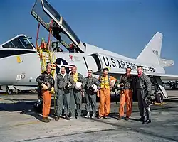 Sept hommes posant en combinaison de vol devant un avion militaire.