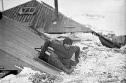 Photographie d'un homme sortant par une ouverture dans le toit d'un petit bâtiment en bois.