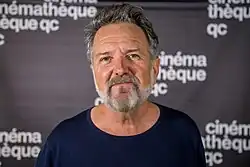 Photo en buste d'un homme souriant aux cheveux et barbe grisonnants et un haut bleu foncé.