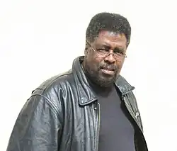 Photo en buste d'un homme noir, portant un blouson en cuir noir et des lunettes.