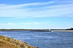 Une étendue d'eau libre de la rivière. Une barge se trouve au centre de l'image, et à l'arrière-plan se trouve une rive boisée. Il n'y a aucun signe d'habitation humaine sur les rives.