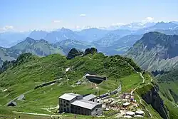 Vue sur un bâtiment-teminus d'une voie de chemin de fer avec à droite, l'arrivée d'une course à pied, et en arrière-plan des montagnes.