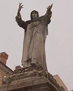 Sculpture d'un personnage sur un piédestal, en toque et capuche levant les bras au ciel