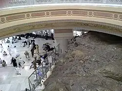 Au premier plan, un gros rocher ; au second plan, des pèlerins en visite, le tout sous le dôme d'un vaste bâtiment