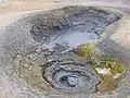 Une mare de boue à Krafla Islande. Notez qu'il s'agit probablement de la même que celle montrée plus en haut, mais prise plus tard, la cavité du bas est d'ailleurs presque tarie et des dépôts de soufre sont clairement visibles autour de la source de jaillissement.