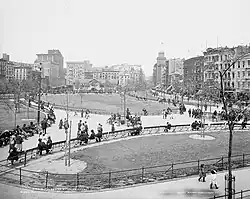 Photographie en noir et blanc de personnes marchant dans les allées d'un parc public, avec en arrière-plan des immeubles et bâtiments hauts.