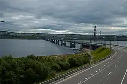 Un pont moderne au centre de l'image enjambe une étendue d'eau sombre. La route allant vers le pont se trouve en bas au premier plan, et en arrière-plan se distingue des collines de l'autre rive vers où file le pont.