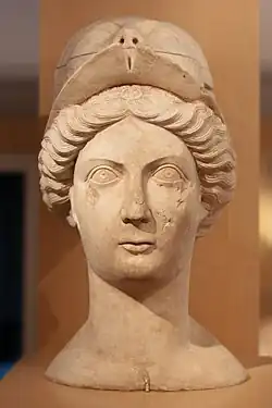 Buste en pierre vu de face, représentant une femme d'allure sévère, aux cheveux ondulés probablement attachés par un chignon. Elle a une petite bouche et des grands yeux et porte un masque glissé sur le haut de sa tête.