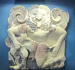 Visage massif de la Gorgone avec la langue pendante, l'œil droit ouvert et le gauche fermé, des ailes en spirale attachées dans le dos, un genou à terre.)