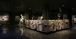 photo d'une salle de musée avec un socle de ±10 x 8 dont les parois verticales sont décorées de jeunes femmes se tenant par l'extrémité de leurs voiles.