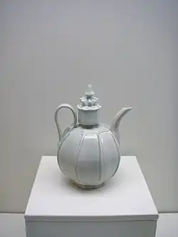 Photographie d'une théière de couleur blanche, avec une large anse et surmontée d'un couvercle travaillé.