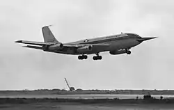 Vue latérale d'un avion volant de la gauche vers le droite de l'image ; une longue perche est visible sur la pointe avant ; le train d'atterrissage est sorti et la piste est visible en-dessous.
