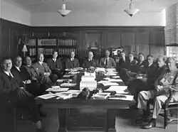 14 hommes en costumes 3 pièces assis autour d'une grande table rectangulaire.