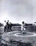 Photographie en noir et blanc d'un homme moustachu lançant un disque, entouré de trois hommes