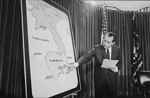 Le président, debout, tient dans sa main gauche plusieurs feuilles et sur une carte de la péninsule indochinoise à sa droite, pointe la frontière sud-orientale du Cambodge.