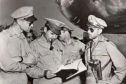 Photos noir et blanc de trois militaires en train d'observer une liasse de papiers que tient celui du centre