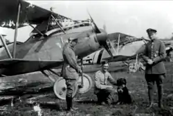 Photo noir et blanc de trois hommes en uniforme, dont l'un est agenouillé près d'un chien. Derrière eux, on peut voir des avions militaires de la Première Guerre mondiale.