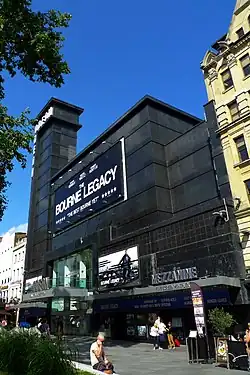 Entrée d'une salle de cinéma dans un édifice noir surmonté d'une tour à gauche.