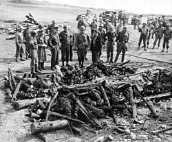 Groupe d'hommes en uniforme devant les restes d'un bûcher où se trouvent les restes calcinés de corps humains.