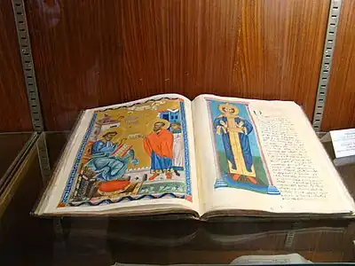 Photographie couleur d'un ouvrage exposé les pages ouvertes. La page de gauche est entièrement peinte, représentant une scène avec plusieurs personnages. La page de droite comporte un texte écrit à la main adossé à une illustration d'un personnage en pied et auréolé.