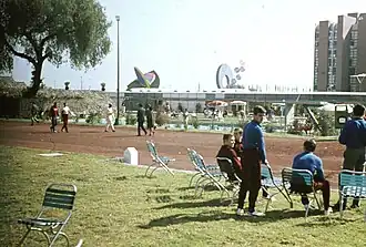 Parc ensoleillé. En fond, le disque solaire et une autre sculpture ainsi qu'une barre d'immeubles. Des personnes sont assises sur des chaises pliantes sur le gazon.