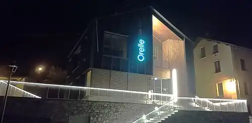Photographie de la face sud de l'office de tourisme (de nuit), avec un éclairage bleu et des maisons dans le parage.