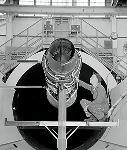photo noir et blanc. Moteur installé sur une plateforme, devant un tunnel de soufflerie. Un technicien, accroupi, l'inspecte.