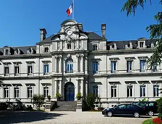 Photo de la façade principale ensoleillée du bâtiment de la préfecture qui s'élève sur trois étages imposants, couverts en ardoises. Au milieu, l'escalier d'une quinzaine de marches mène à un portail fermé à double vantail, entouré de chaque côté de deux colonnes. Au premier étage, autant de colonnes encadrent une grande fenêtre. Au-dessus, les colonnes ont fait place à des pilastres qui encadrent une petite fenêtre. Il est surmonté d'un fronton au milieu duquel se trouve une horloge. Sur le toit, un drapeau français flotte au sommet d'un mat.