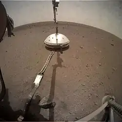 Photo montrant la cloche recouvrant le sismomètre posé à la surface de Mars relié par un câble à l'atterrisseur et toujours accroché au câble qui l'a déposé.