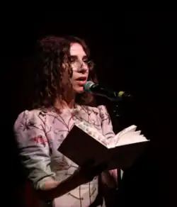 Image d'une femme brune avec des lunettes portant un haut gris brodé et lisant un livre en parlant dans un micro