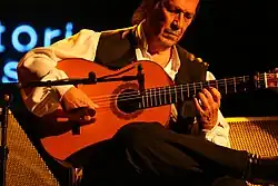 Photographie en plan rapproché d'un homme dans la maturité, assis, jouant de la guitare les yeux mi-clos, la guitare posée sur son genou droit, jambe repliée posée sur genou gauche, dans la posture habituelle pour la guitare flamenca.