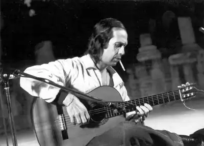Photographie en noir et blanc d'un homme assis jouant de la guitare, une cigarette aux lèvres.
