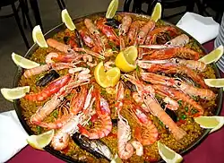 Photographie couleur représentant un plat garni. Le riz safrané garni de petits pois et de mollusques et surmonté de langoustines, moules et quartiers de citron.