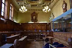 salle lambrissée avec de nombreux sièges, un box vitré à droite et un plafond peint à caissons