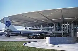Photo d'un avion blanc avec des bandes bleues stationné sur le tarmac d'un aéroport. Le terminal, visible en arrière-plan, a une architecture moderne avec des baies vitrées et un toit large et plat, soutenu par des colonnes.
